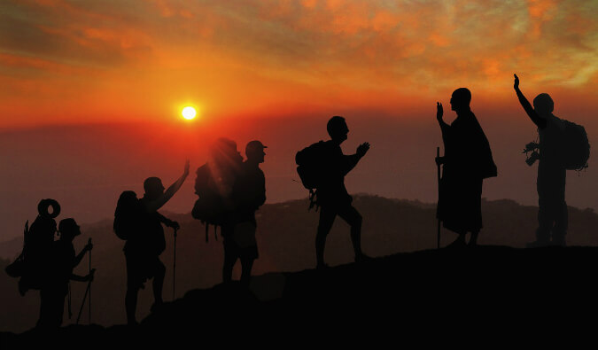 4 hikers with backpacks walking up a mountain being greeted by 2 people at the top all in silhouette at red sunset