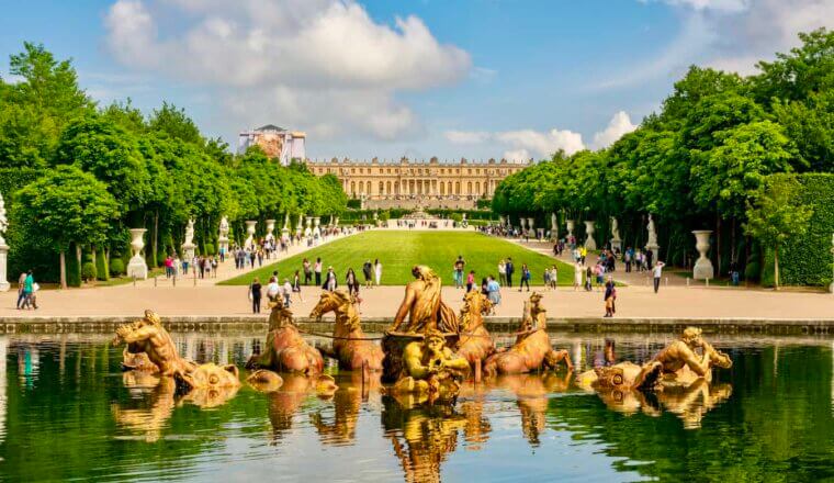 The statues and beautiful facade of the Palace of Versailles in France with people exploring the grounds in the distance