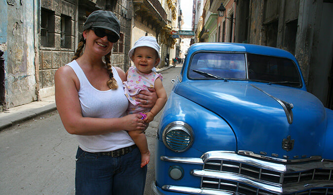 woman holding a baby in Cuba next to a blue classic car