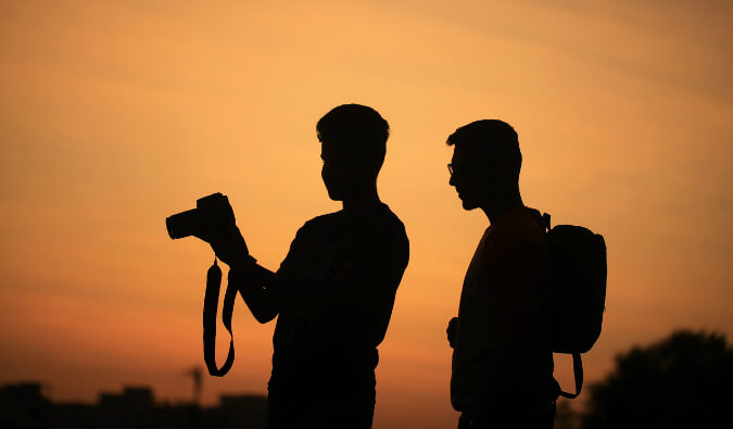 two men looking at photos on a DSLR camera silhouetted at sunset