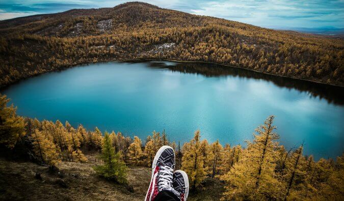 feet wearing sneakers front centre above a beautiful lake surrounded by trees and greenery in the background