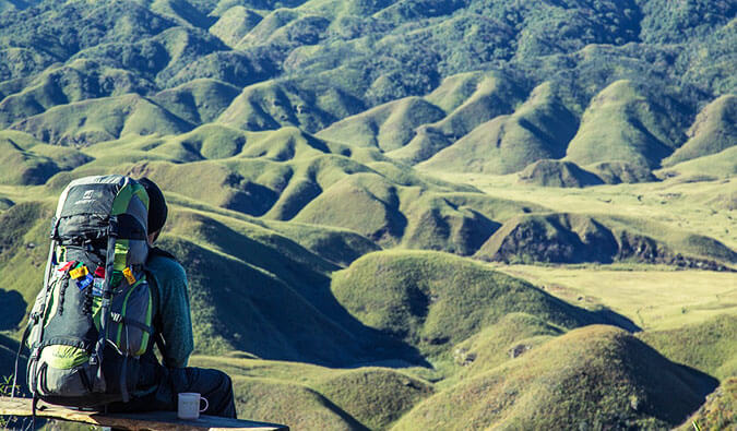 Man sat on a wall with a backpack on and a mug next to him looking out to the view of green hills