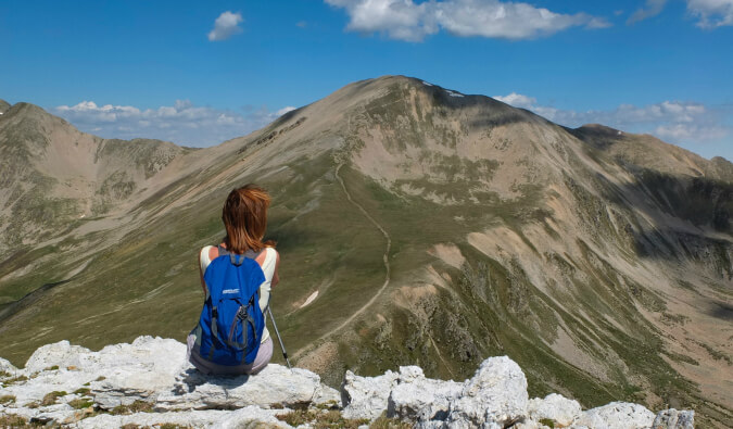 woman with hiking stick and backpack sat facing away from camera looking out to the mountains