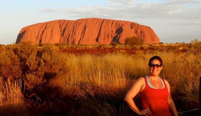 woman in foreground wearing sunglasses smiling tall grass and large rock in background