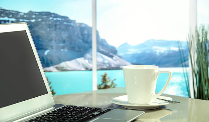 laptop and coffee cup and saucer on a table next to a window with a view of a lake and mountains in the background
