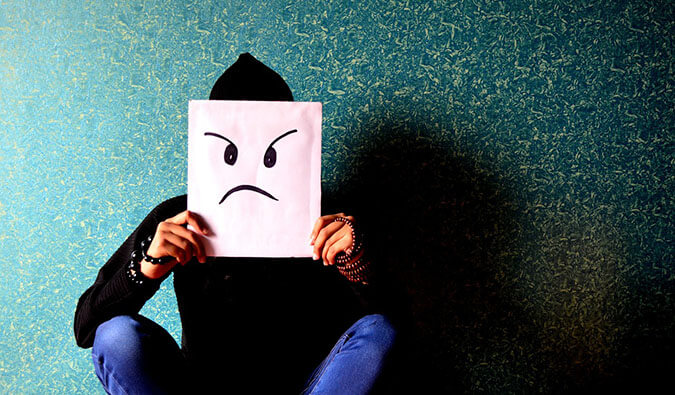 man sat in front of a green patterned wall holding a white square with a frowning face drawn on in front of his face