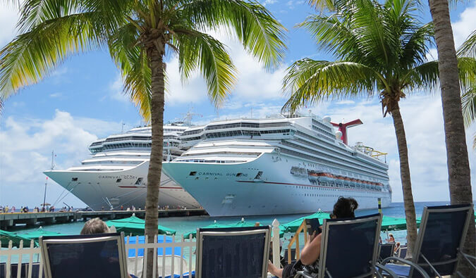 Two cruise ships docked in the Caribbean. People in the foreground sunbathing on sun beds reading