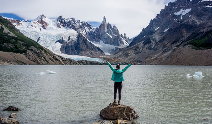 Woman in a green jacket standing on a rock in a lake surrounded by snowcapped mountains