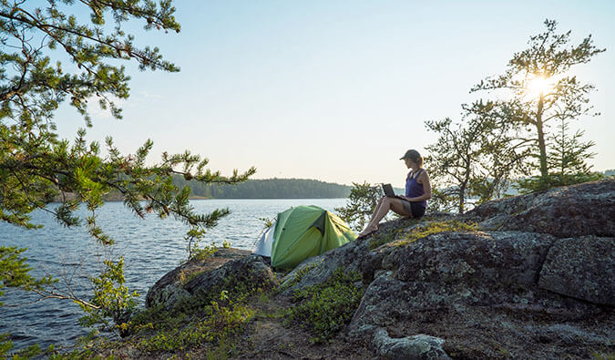 Woman sat reading a book close to her tent which is pitched on the bank of a river