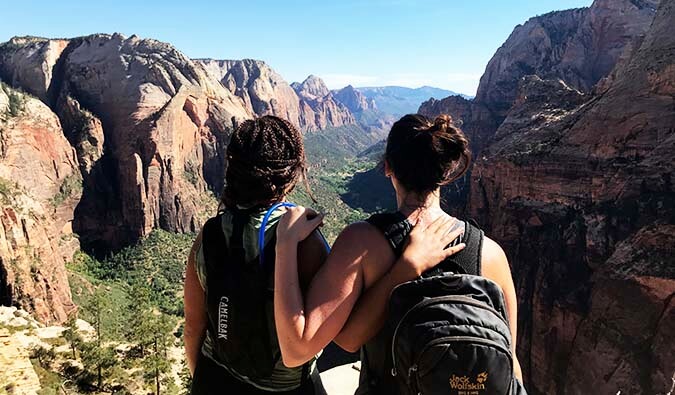 Two girls with their arms around each other wearing backpacks looking out to a view of mountains