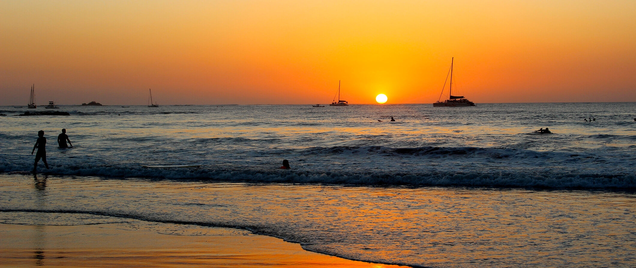 A beach in Tamarindo at sunset during the golden hour. People swimming in the sea, sailboats in the distance