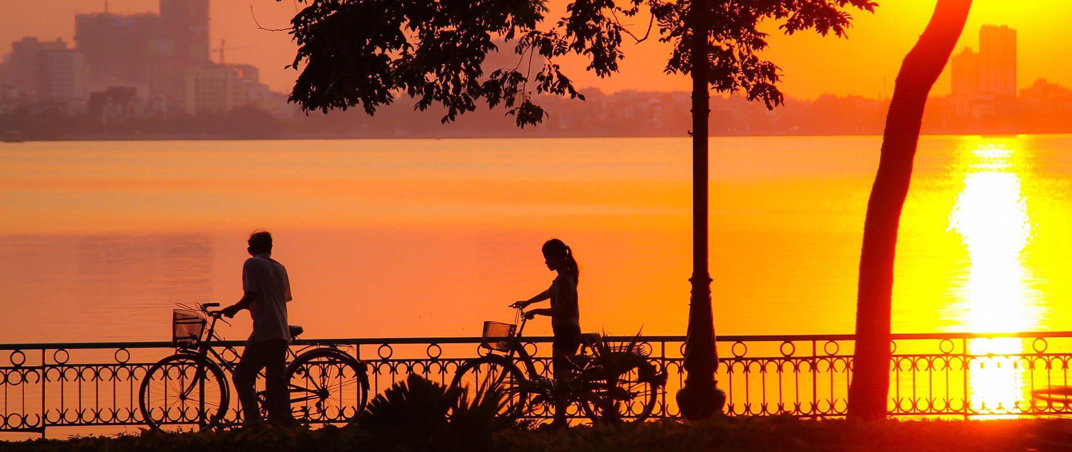 two people walking with bicycles across a bridge silhouetted in the sunset.