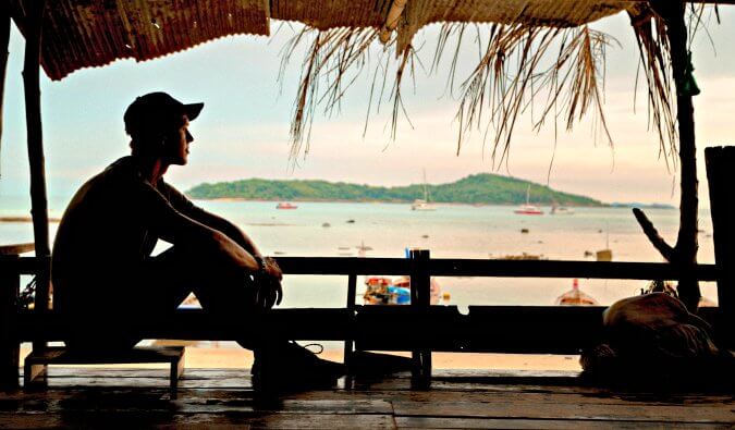 man part silhouetted sitting in a wooden hut with his backpack in foreground. In the background beach boats and sea