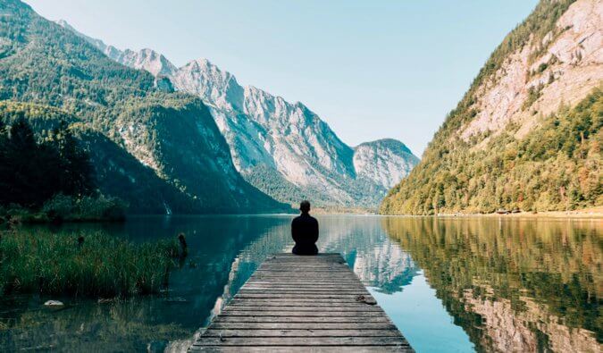 man sat on the end of a pontoon wearing black surrounded by a lake and mountains