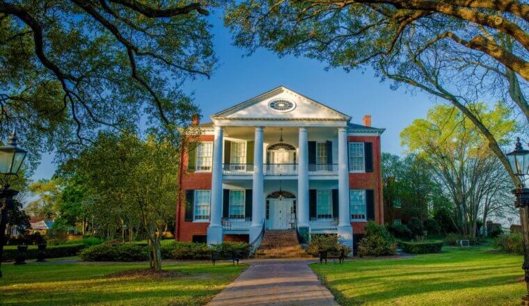 A pathway leading up to the historic Rosalie mansion, with white pillars and a brick façade with black wooden shutters, surrounded by lush trees in Natchez, Mississippi, USA