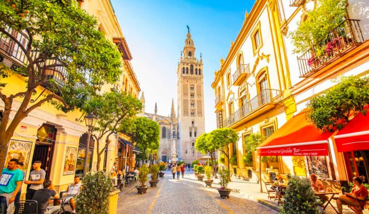 People walking along a quiet, narrow street in sunny Sevilla, Spain with a church tower in the distance