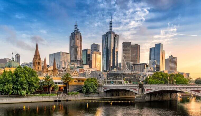 The towering skyline of Melbourne, Australia with trees and a bridge in the foreground near the river