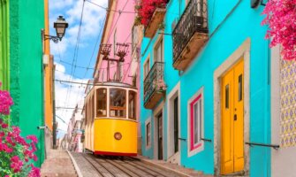 Historic yellow tram on a street going down a hill lined with colorful buildings and flowers on the balconies in Lisbon, Portugal