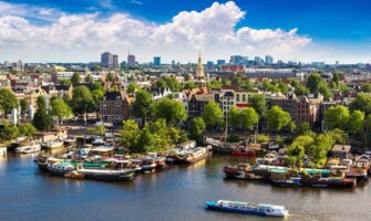 Panoramic view of the Amsterdam skyline with boats docked and floating along a river in the foreground