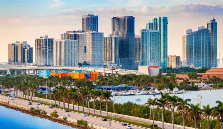 The downtown skyline of Miami at sunset, with skyscrapers in the background and a palm-tree-lined waterfront boulevard in the foreground